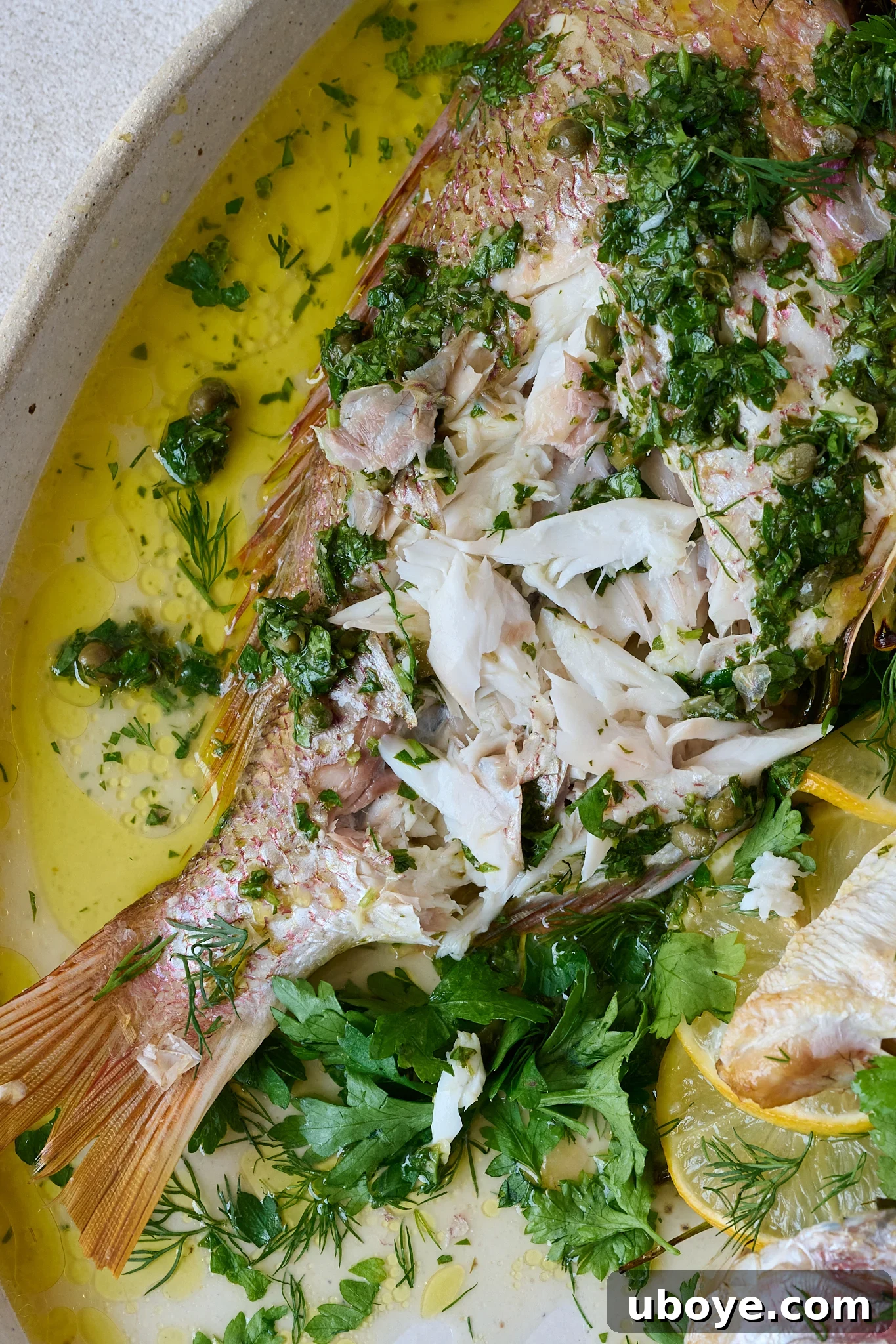 A close-up of the whole baked fish with salsa verde on a large white platter, showing the flaky white flesh exposed after a portion has been served, beautifully surrounded by fresh herbs and lemon slices.