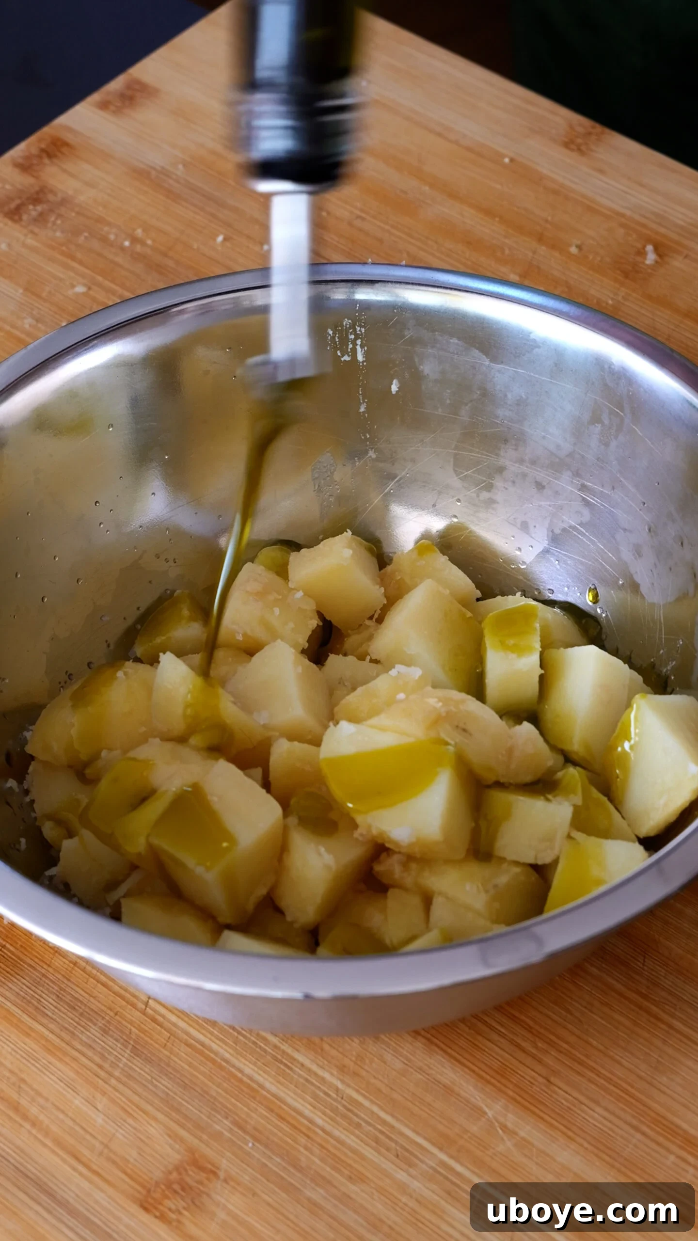 Boiled potatoes are in stainless steel bowl and being dressed with olive oil