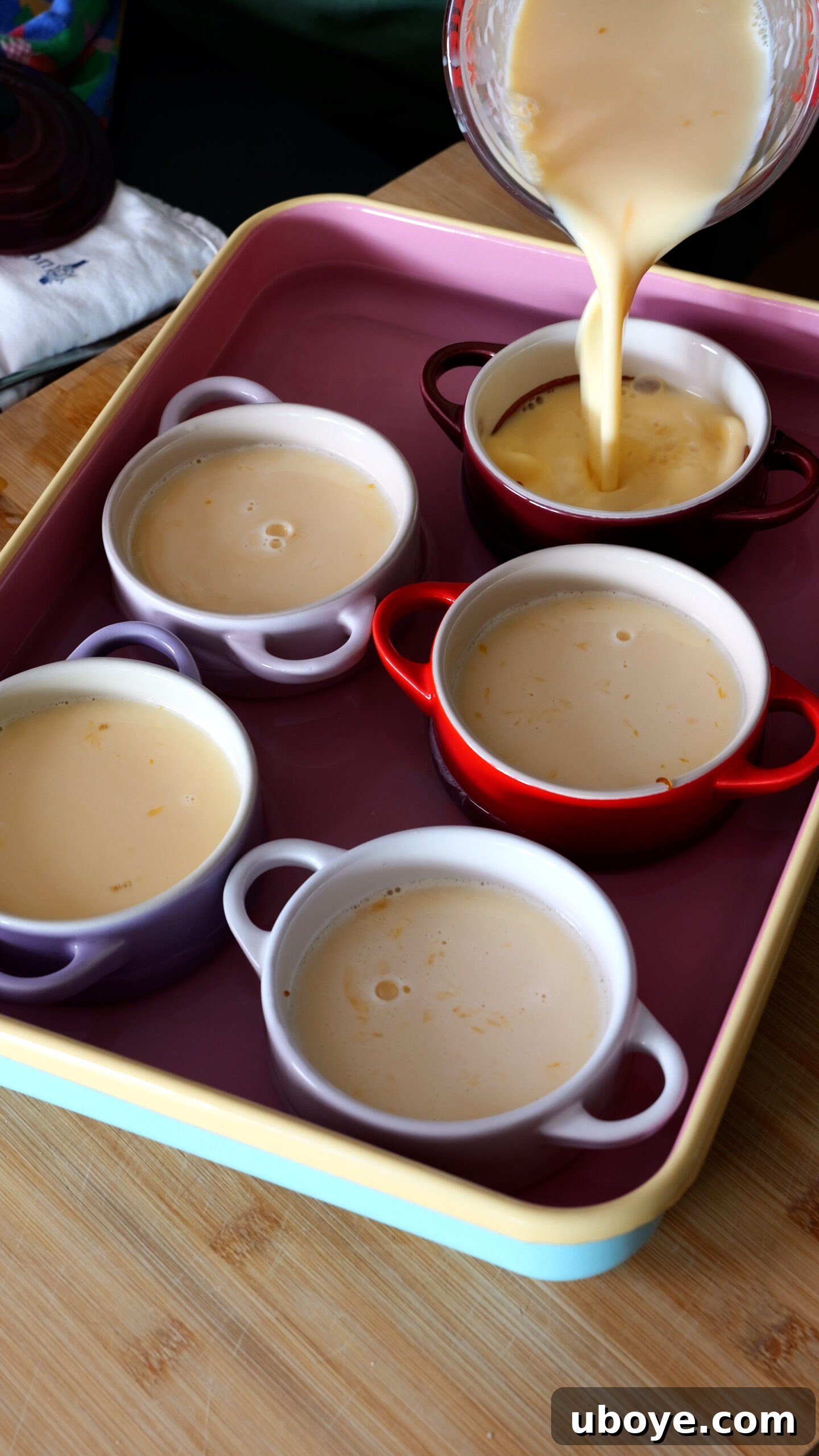 Classic Spanish Flan Recipe: Smooth Caramel Custard You’ll Love 7 five ramekins are in large pink baking tray and being filled with custard.